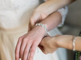 An intricate diamond bracelet is secured onto a woman's wrist by another person assisting her as she holds out her arm.