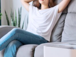 A young woman sits on a gray couch with her eyes closed and hands behind her head. A white air purifier is next to the sofa.