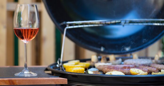A glass of red wine in a traditional glass rests next to a charcoal grill. Corn and some meat are on the grill.