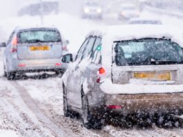 Two cars driving on a slippery snow covered road as more snow covers the cars and road. There are other cars in the distance.