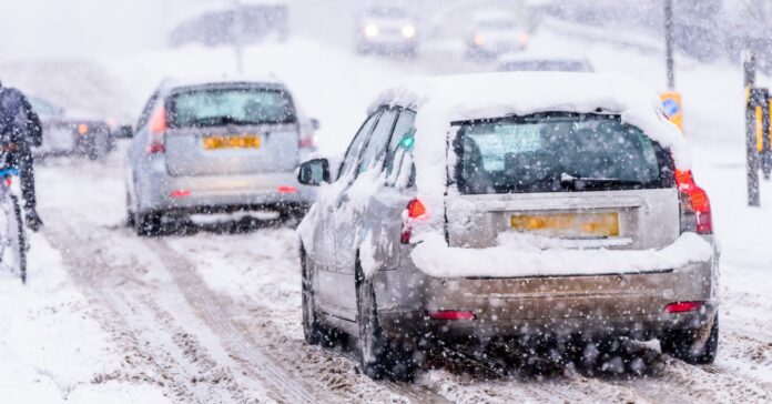 Two cars driving on a slippery snow covered road as more snow covers the cars and road. There are other cars in the distance.