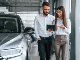 A man and a woman wearing white shirts stand side by side while the man holds a tablet next to a silver car.