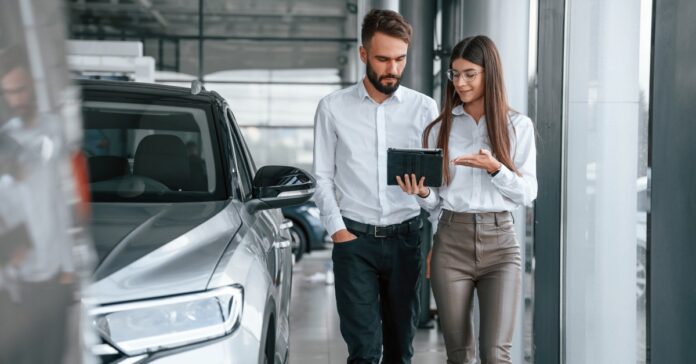 A man and a woman wearing white shirts stand side by side while the man holds a tablet next to a silver car.
