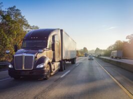 A black semi-truck drives down a highway with other cars in the distance behind it under a clear blue sky.