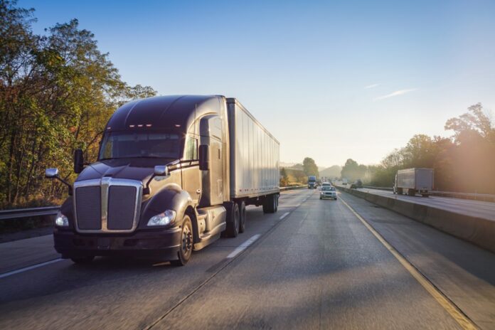 A black semi-truck drives down a highway with other cars in the distance behind it under a clear blue sky.