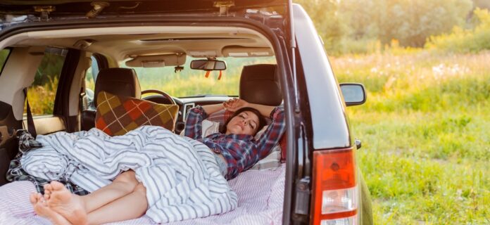 A camper sleeps comfortably in her car’s trunk under a striped blanket, next to a brown plaid pillow.