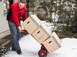 A man in a red jacket using a hand truck to move three cardboard boxes stacked on top of each other through the snow.