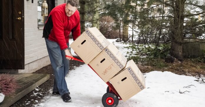 A man in a red jacket using a hand truck to move three cardboard boxes stacked on top of each other through the snow.