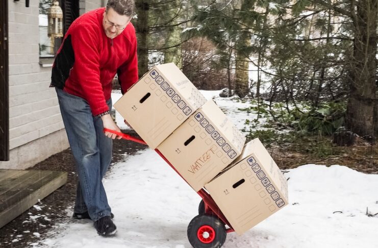 A man in a red jacket using a hand truck to move three cardboard boxes stacked on top of each other through the snow.