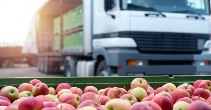 The top of a green crate filled with red-and-yellow apples with a large loading truck in the background with an open container.