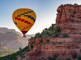 A yellow hot air balloon floats next to some red rock mountains in Sedona, Arizona at sunrise.