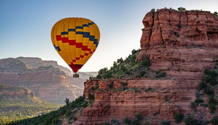 A yellow hot air balloon floats next to some red rock mountains in Sedona, Arizona at sunrise.