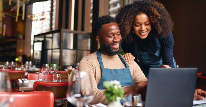 A woman in a navy sweater points to a laptop screen as a seated man in a green apron smiles beside her.