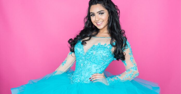 A teenager with loosely curled hair stands with her hand on her hip, wearing a vibrant cyan ballgown with sheer sleeves.
