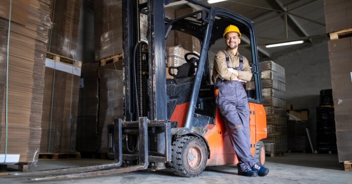 An employee wearing a yellow hard hat is smiling and leaning against a parked forklift. The employee crosses their arms.