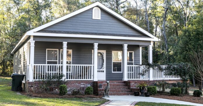 A manufactured home has gray siding and a red brick front porch with white pillars. A concrete walkway leads to the porch.