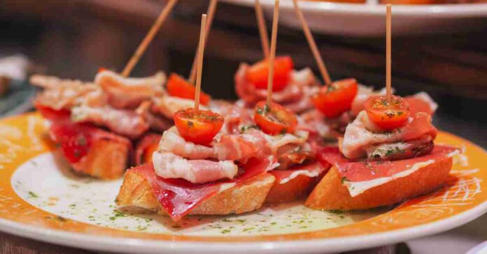 A plate of pintxos from San Sebastián, Spain. There are pieces of toasted bread with cured meats and a cherry tomato speared on top.