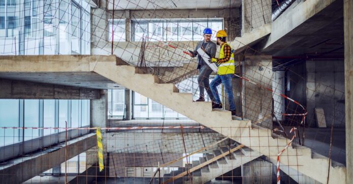 Two people wearing hard hats walking up a large staircase in a building that is in the middle of construction.