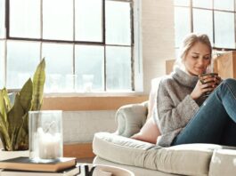 A smiling woman wearing a sweater and jeans sits on a cream-colored couch holding a mug with both hands.