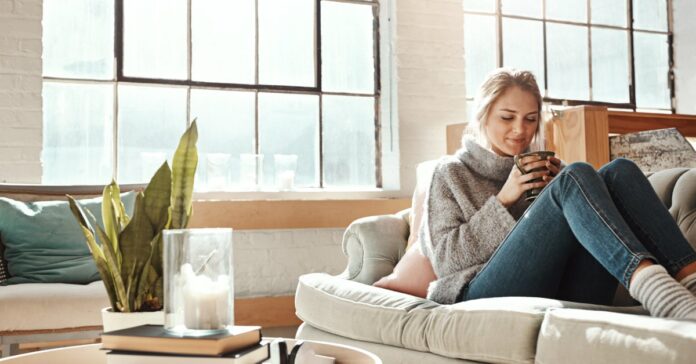 A smiling woman wearing a sweater and jeans sits on a cream-colored couch holding a mug with both hands.
