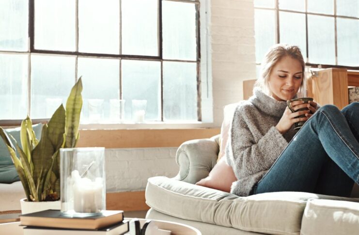 A smiling woman wearing a sweater and jeans sits on a cream-colored couch holding a mug with both hands.