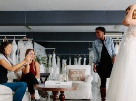 How To Make Wedding Dress Shopping More Fun A bride-to-be in a wedding dress and veil smiles as three women watch her try it on in a bridal salon.