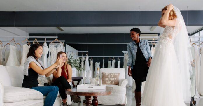 A bride-to-be in a wedding dress and veil smiles as three women watch her try it on in a bridal salon.