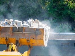 A bulldozer's crane lifting a heavy load of white bricks and other debris high in the air near a wall by a forest.