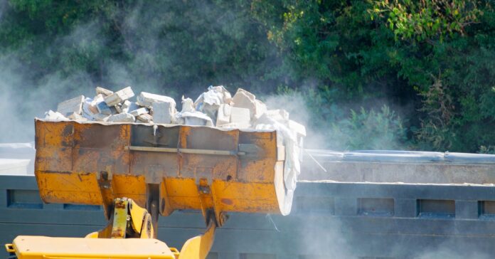 A bulldozer's crane lifting a heavy load of white bricks and other debris high in the air near a wall by a forest.