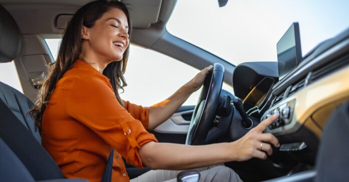 A woman sits in the driver's seat of a car and smiles while she touches a dial on the car's stereo system.