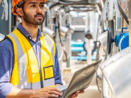 How Solvent Recycling Improves Business Sustainability Man in orange hard hat and yellow safety vest holds laptop while standing among industrial pipes and equipment outdoors.