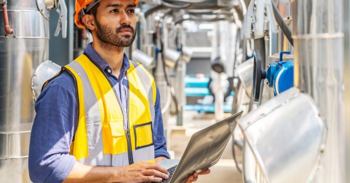 Man in orange hard hat and yellow safety vest holds laptop while standing among industrial pipes and equipment outdoors.