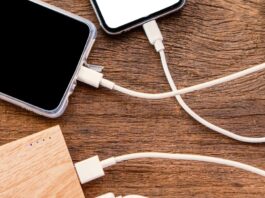 Four white charging cables attached to two smartphones and a light brown power bank sitting on a wooden table.