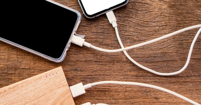 Four white charging cables attached to two smartphones and a light brown power bank sitting on a wooden table.