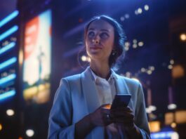 A smiling professionally dressed woman looks up from her smart phone to gaze at her neon-lit nighttime urban surroundings.