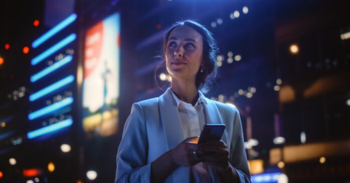 A smiling professionally dressed woman looks up from her smart phone to gaze at her neon-lit nighttime urban surroundings.