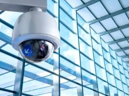 Innovative Security Solutions for Modern Businesses A white and black security camera is mounted high on a wall with floor-to-ceiling windows spanning one wall.
