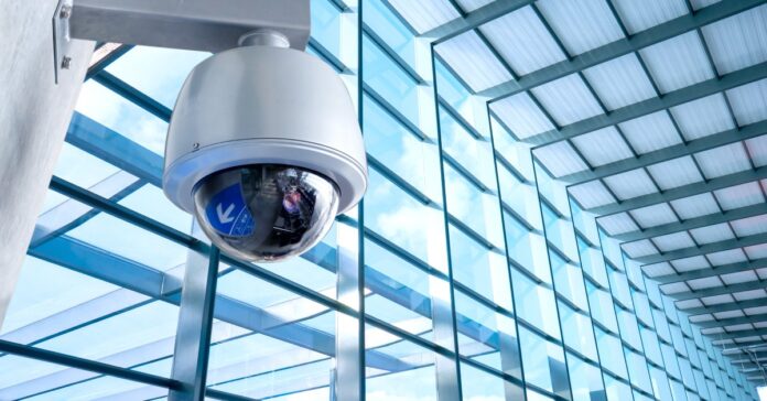 A white and black security camera is mounted high on a wall with floor-to-ceiling windows spanning one wall.