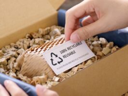A person holds a slip of paper that reads 100% Recyclable, Reusable. The paper sits inside a box filled with eco-friendly paper-based packing materials.