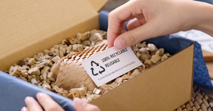 A person holds a slip of paper that reads 100% Recyclable, Reusable. The paper sits inside a box filled with eco-friendly paper-based packing materials.