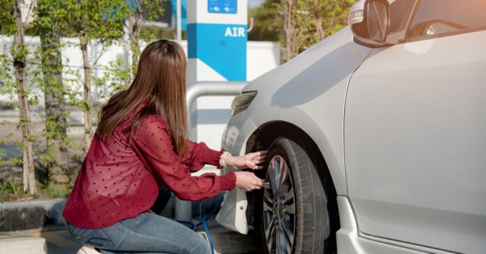 A woman in a red shirt and blue jeans airs up the front tire of her white car at an air pump in a gas station parking lot.