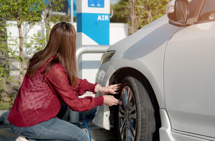 A woman in a red shirt and blue jeans airs up the front tire of her white car at an air pump in a gas station parking lot.