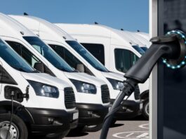 Several white electric vans are parked in a row in a lot, with an electric vehicle charging station in front of them.