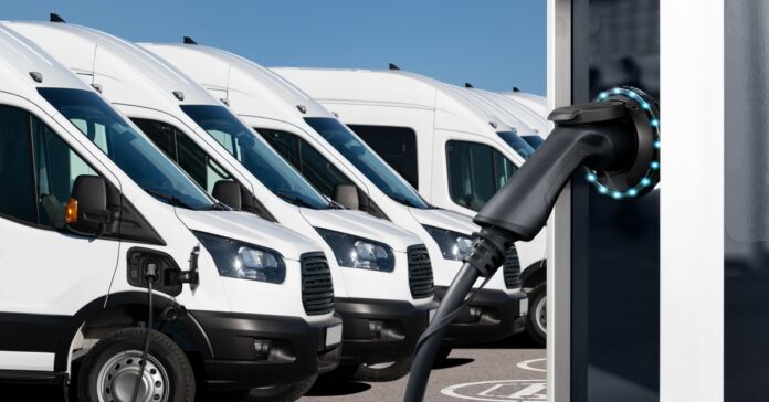 Several white electric vans are parked in a row in a lot, with an electric vehicle charging station in front of them.
