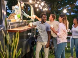 Five people excitedly crowd around a food truck outside. Bright lights attached to the truck illuminate the space.