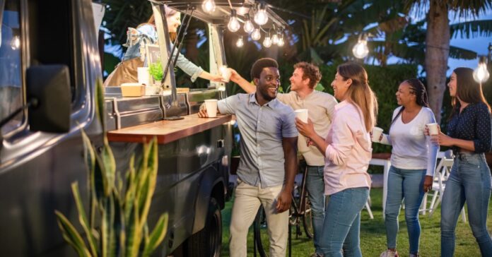 Five people excitedly crowd around a food truck outside. Bright lights attached to the truck illuminate the space.