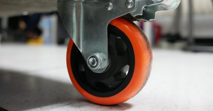 A close-up of a bright orange polyurethane caster wheel on a trolley. The wheel shows some dirt and wear.