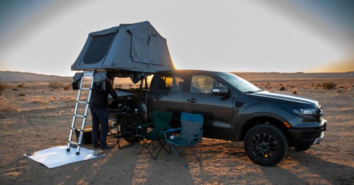 A man sets up a truck bed tent with a ladder in a desert at sunset, with camping chairs and a grill next to the truck.