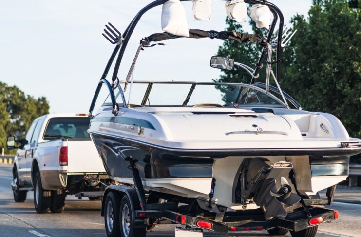 A white four-door truck tows a large white motorboat across an interstate bridge with trees lining the railing.