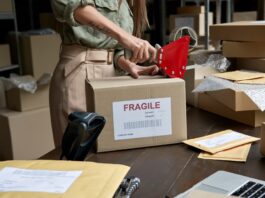 A woman in a green shirt tapes a box labeled fragile amid packing supplies in a room lined with shelves of stored boxes.